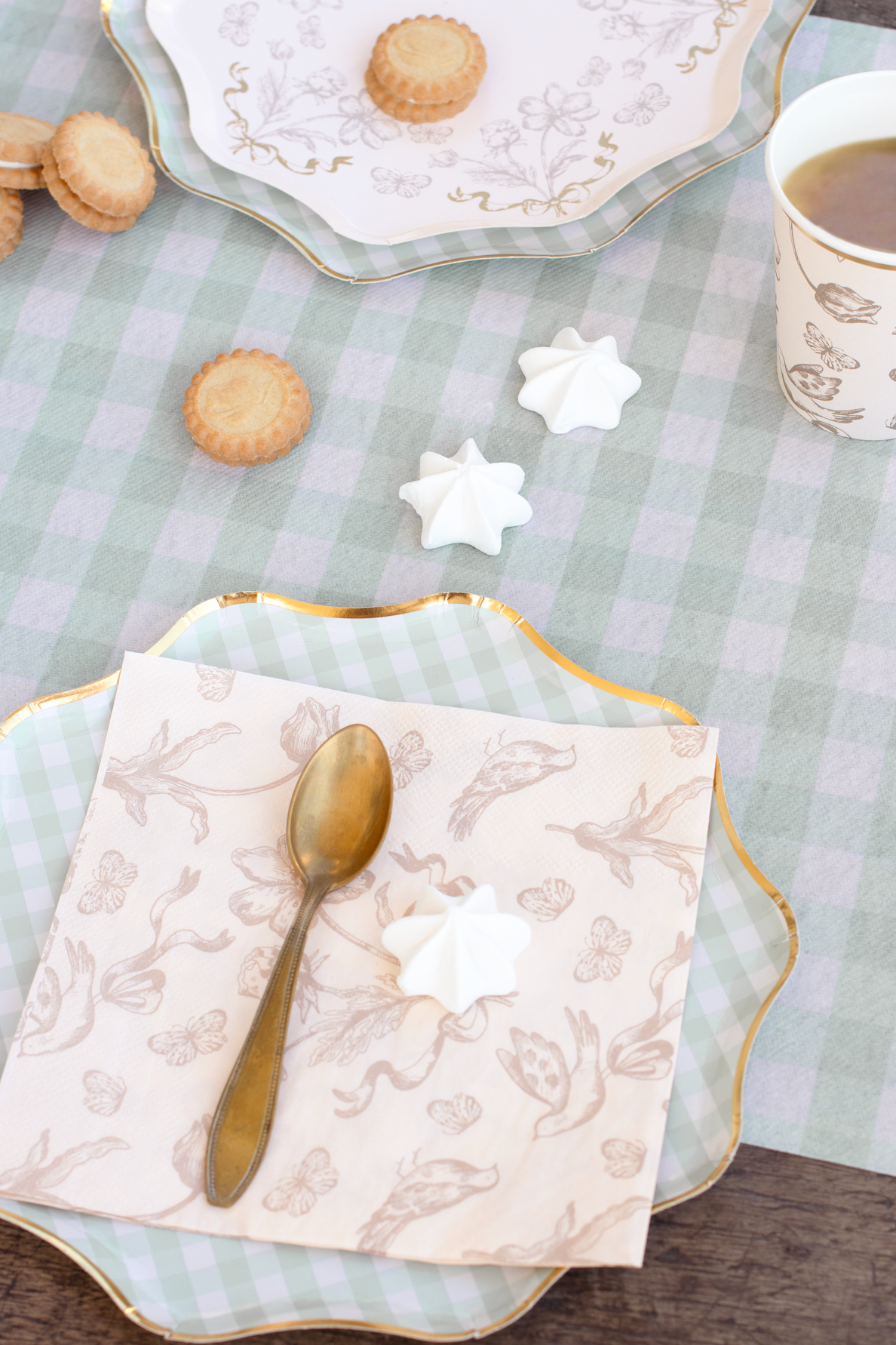 Decorative table setting with a teacup, cookies, and a spoon on a checkered tablecloth.