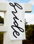 White towel with 'Bride' in black cursive text draped over a white railing outdoors.