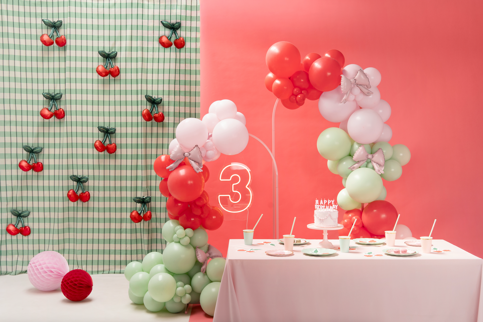 Children's birthday party setup with balloons, cake, and decorations against a pink and green checkered wall.