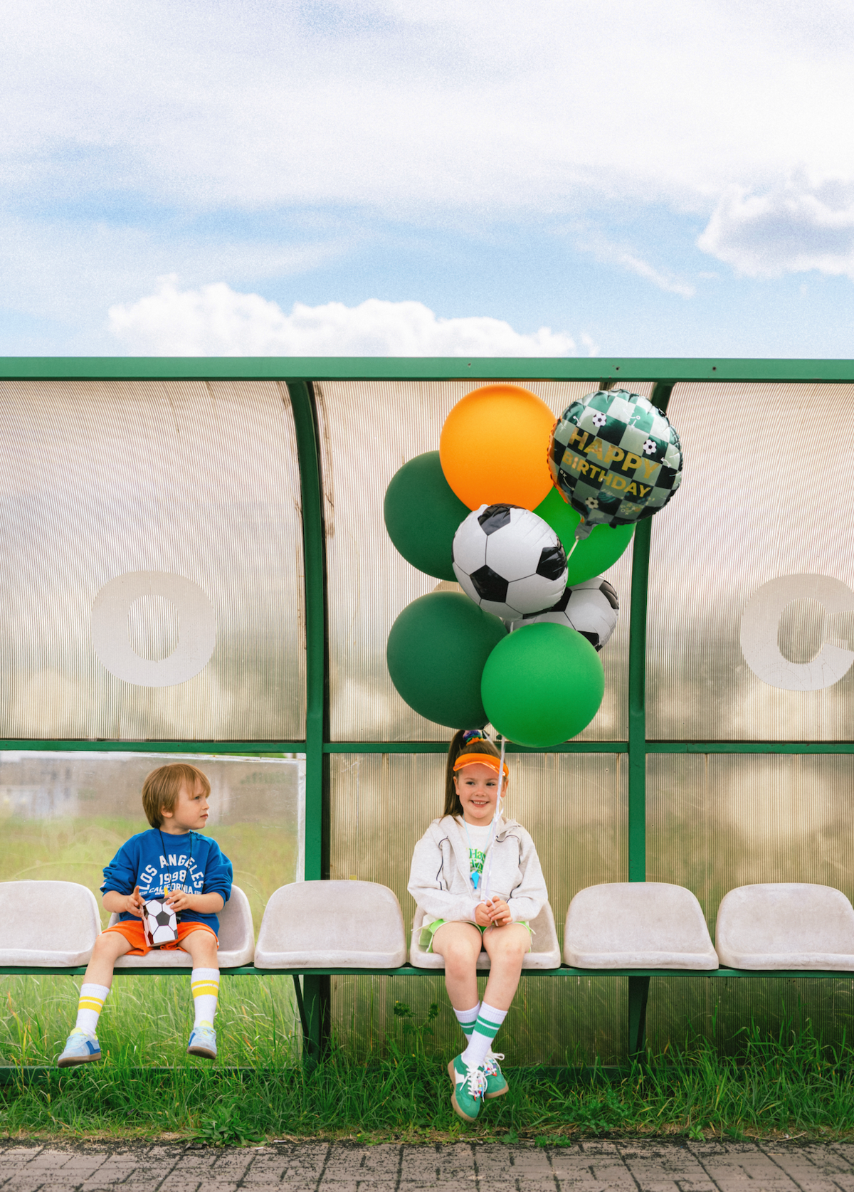Two children sitting on a bench with sports-themed balloons in front of a bus stop.
