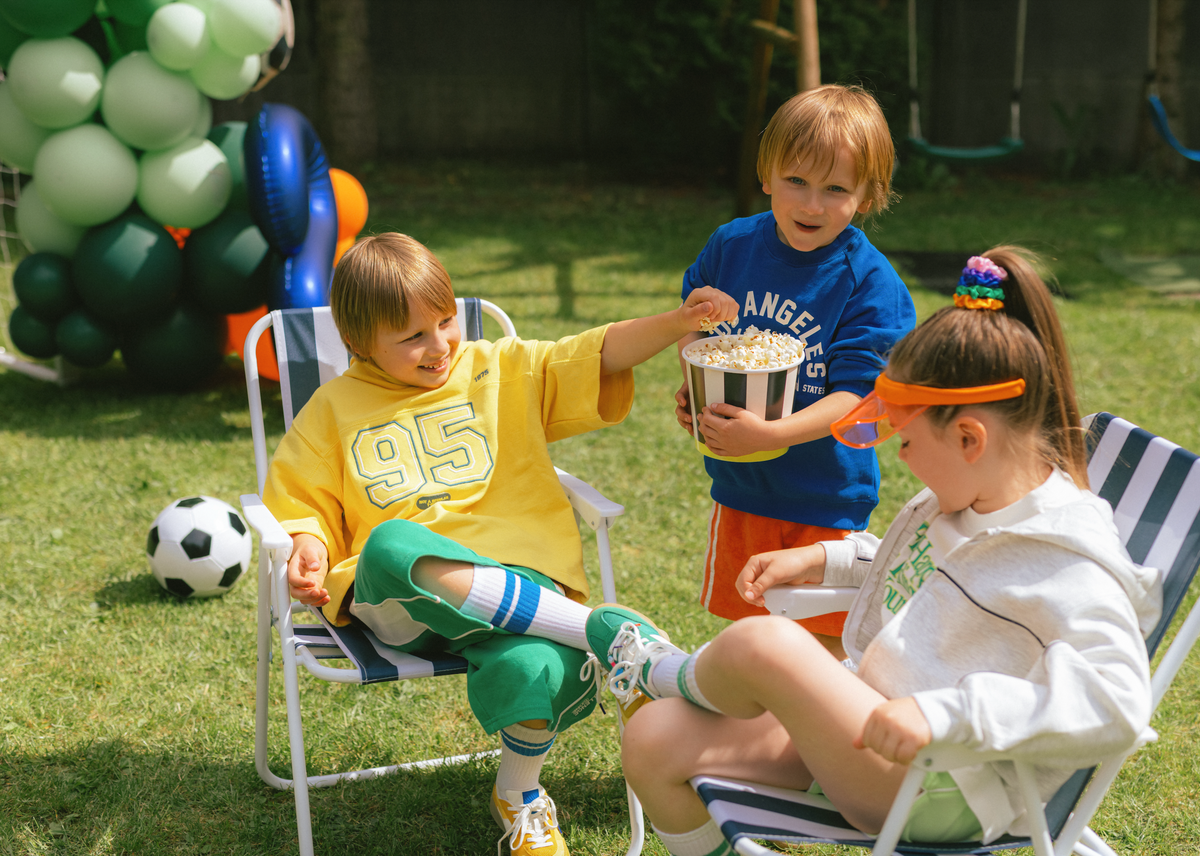 Children sitting on lawn chairs outdoors with balloons and sports balls in the background