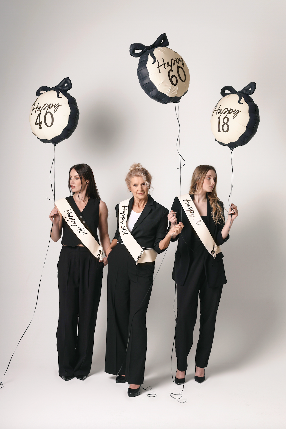 Three women holding balloons with numbers and &#39;Happy Birthday&#39; sashes against a white background