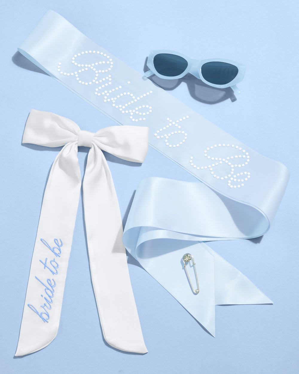 White sash with 'bride to be' text, sunglasses, and a brooch on a light blue background