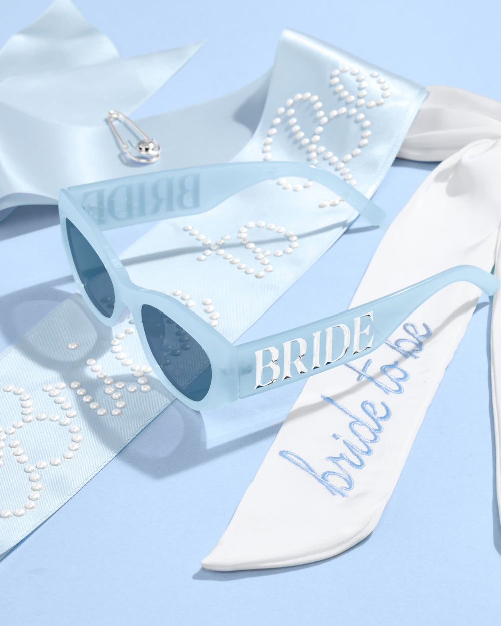 White sash with 'bride to be' text, sunglasses, and a brooch on a light blue background