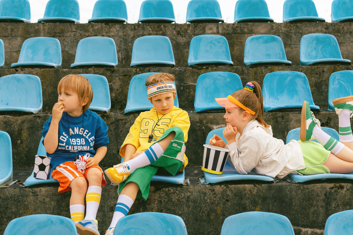 Three children sitting on blue stadium seats with sports gear and snacks.