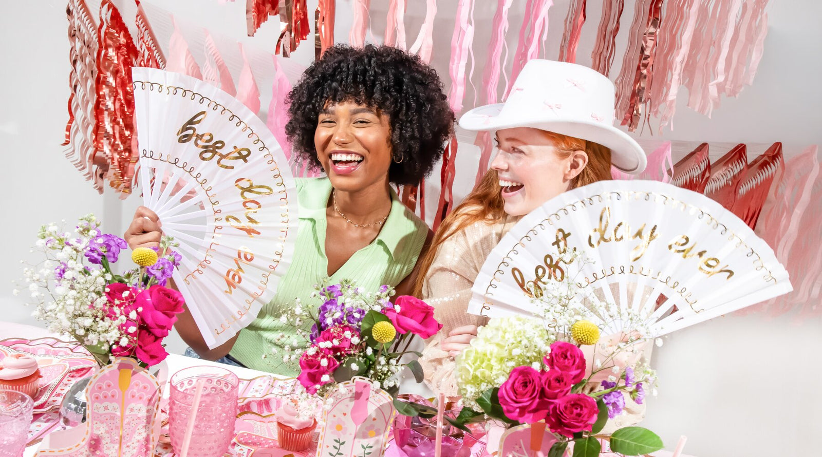 Two women laughing at a pink and white bachelorette party table, each holding a white folding fan with gold script that reads 'honey, let's eat dinner'. The scene is decorated with pink and red tinsel, flowers, and pink cups.