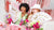 Two women laughing at a pink and white bachelorette party table, each holding a white folding fan with gold script that reads 'honey, let's eat dinner'. The scene is decorated with pink and red tinsel, flowers, and pink cups.