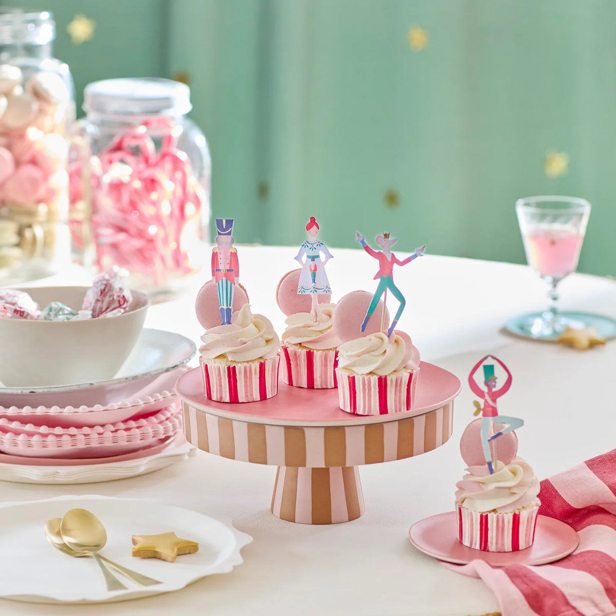 Cupcakes with dancer toppers on a pink striped cake stand with candy and a glass in the background.