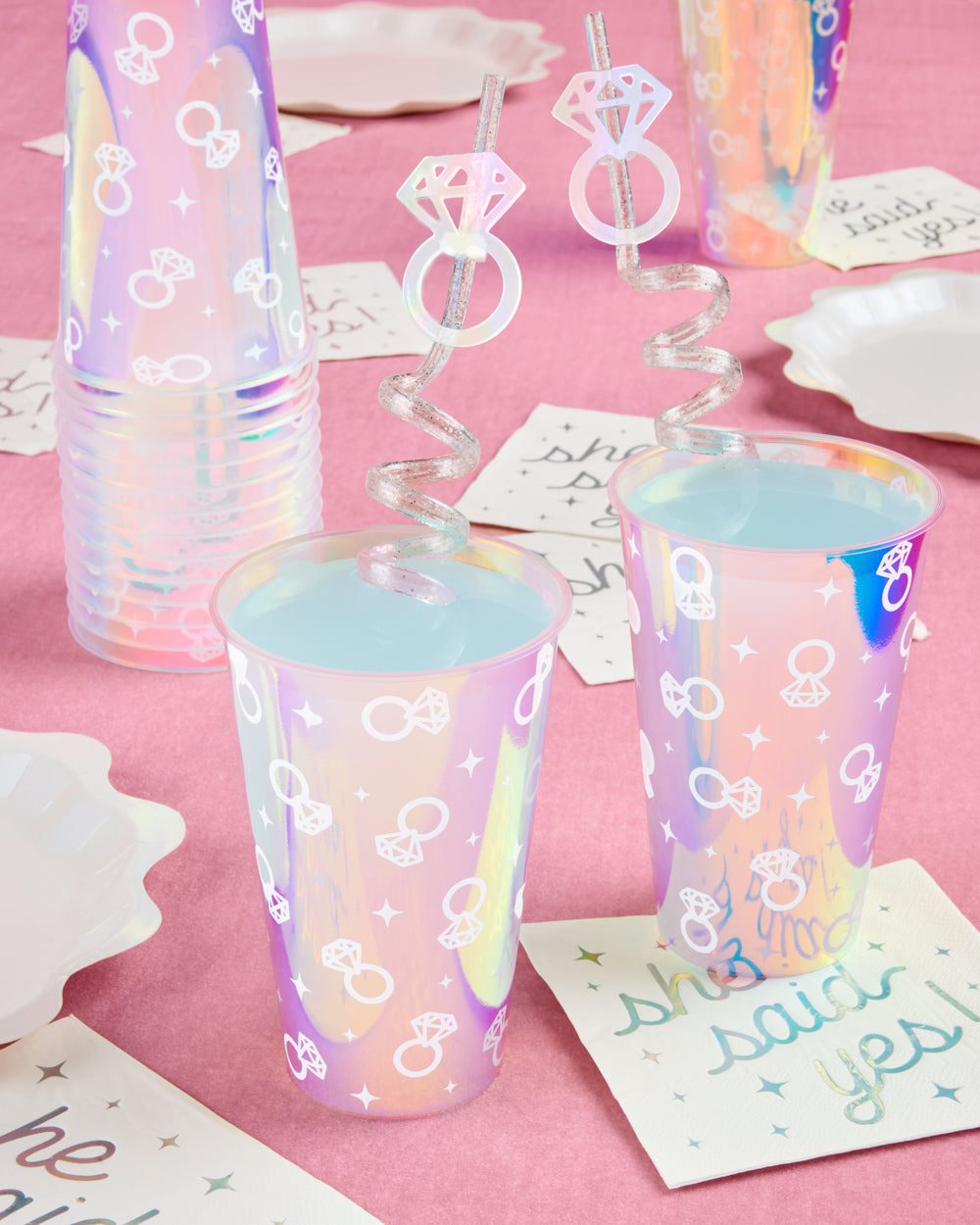 Iridescent party cups with bow and ring designs on a pink surface.